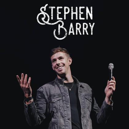 Stephen Barry, singer holds out both hands. Left hand holds a microphone. He is smiling wearing a grey denim jacket over a black T-shirt . Background is black.